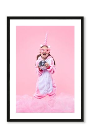 Happy girl in pink unicorn onesie laughing, wearing oversized pink glasses and holding disco ball against pink celebratory background. Concept of childhood, fantasy, dreams, imagination, party