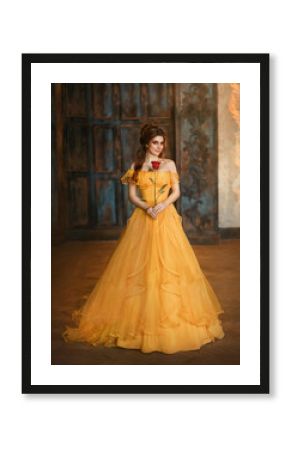 Girl beauty fantasy princess in yellow long historical, medieval silk dress holding flower red rose in her hands. Background of old gothic castle room. Fairy tale bewitched queen. Happy woman smiles