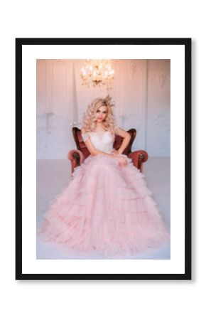 elegant beautiful young princess girl. Blonde queen woman sitting in old medieval vintage armchair. Golden crown pink vintage long dress. Backdrop shines crystal chandelier white interior classic room