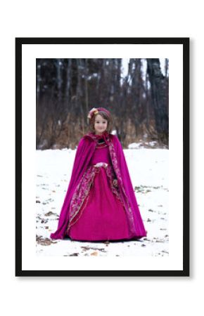Cute toddler girl in a beautiful velvet royal princess outfit with a cape and a long dress. Child is standing in the snowy forest, Halloween costume.