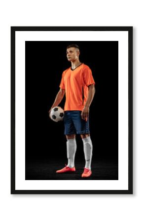 Young male soccer player in orange-blue football kit posing with ball isolated on dark background. Concept of sport, goals, competition, male hobby, occupations