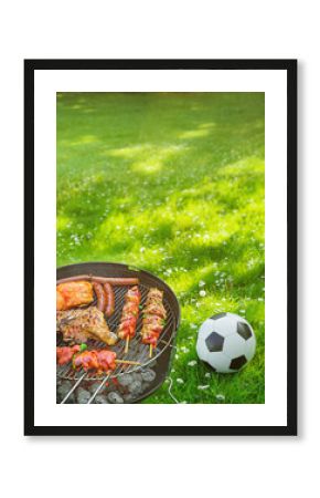  Delicious grilled meat with vegetables sizzling over the coals on barbecue with a soccer ball on the green meadow in the background. Poster.