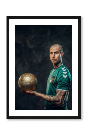 Bold and handsome soccer player posing for a photoshot in a dark studio, wearing green professional sportswear, and holding a golden soccer ball looking serious and focused