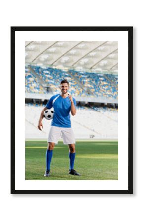 happy professional soccer player in blue and white uniform with ball showing yes gesture on football pitch at stadium