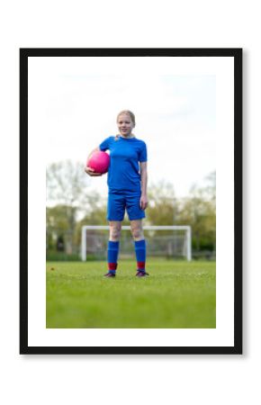 A smiling young girl in blue sportswear holds a pink soccer ball on a grassy field.