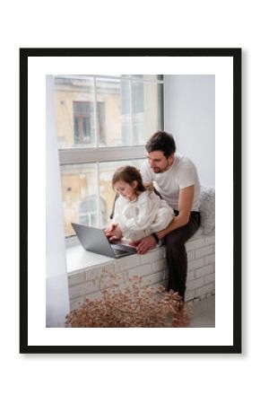 Father and daughter are playing on laptop, sitting on wooden, white windowsill near the window