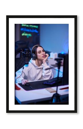 Happy woman putting on headphones and playing computer game. Female gamer in glasses sitting and looking at computer monitor.