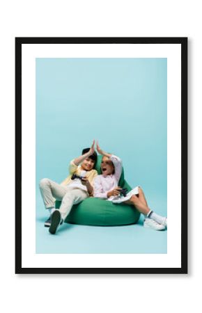 KYIV, UKRAINE - JULY 2, 2021: Multiethnic kids holding joysticks and giving high five on beanbag chair on blue background.