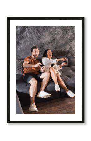 KYIV, UKRAINE - JULY 26, 2022: happy multiethnic friends playing video game and sitting on grey couch