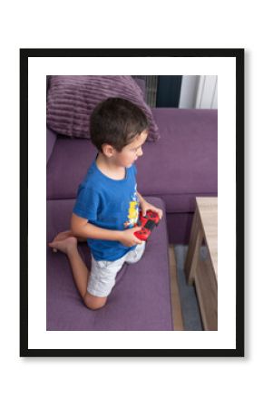 Young man playing video games enthusiastically on a purple sofa A boy in a blue t-shirt and gray shorts is kneeling on a purple sofa, excitedly holding a red game controller.