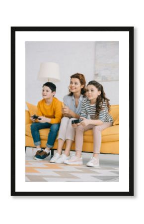 happy mother sitting on yellow sofa near adorable daughter  and son playing video game with joysticks