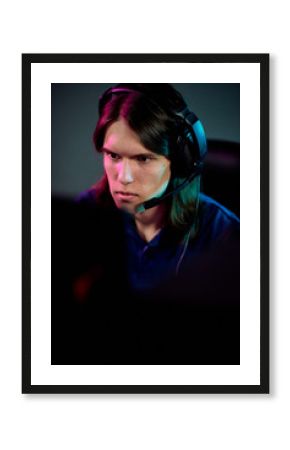 Focused guy with long hair using headset with microphone for communication with network gamers while playing computer game