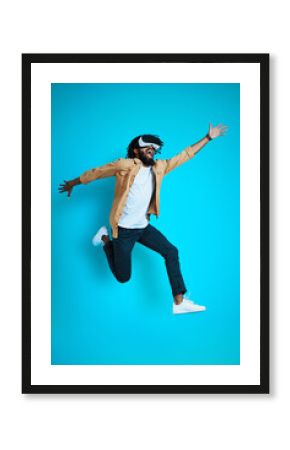 Full length of playful young African man wearing virtual reality glasses while hovering against blue background