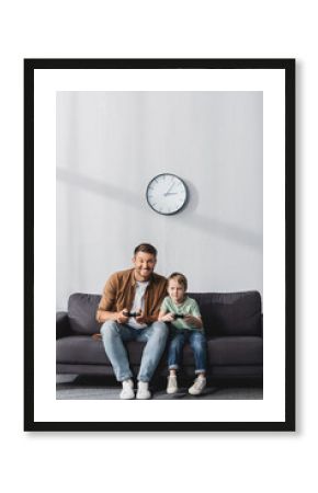 KYIV, UKRAINE - JUNE 9, 2020: happy father and son sitting on sofa and playing video game with joysticks