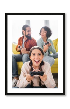 KYIV, UKRAINE - NOVEMBER 16, 2020: excited hispanic girl playing video game near parents holding joysticks on blurred background