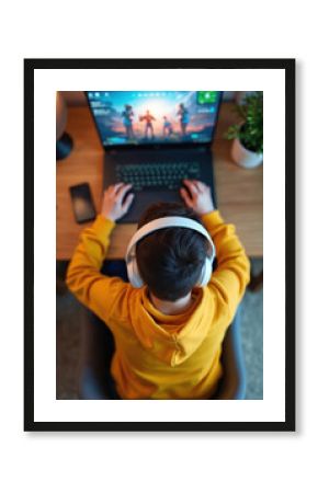 Young man wearing white headphones plays computer games on laptop. Top view of gamer at home during quarantine. Sits, plays online game with immersed concentration, using technology for