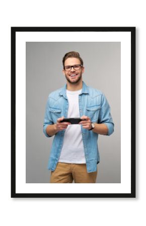 Young handcome man playing with portable video game standing over grey background