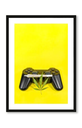 Video games and cannabis: game controller with marijuana leaf and big cannabis joint isolate on yellow background with copy space top.