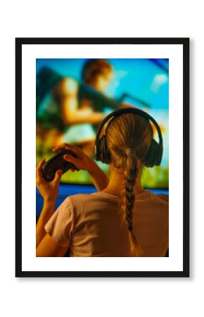 Young woman shooter plays an exciting video game. In front of her is a large computer monitor, a joystick in her hands, headphones on her head. Close-up. Shooting from the back.