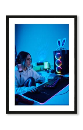 Happy woman putting on headphones and playing computer game. Female gamer in glasses sitting and looking at computer monitor.