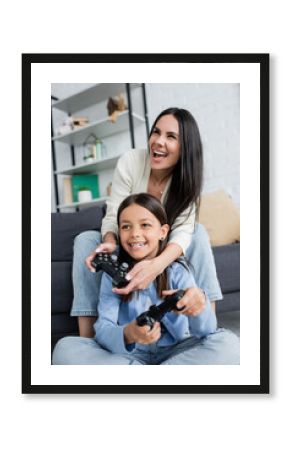 KYIV, UKRAINE - MAY 23, 2022: excited nanny and child playing video game with joysticks at home.