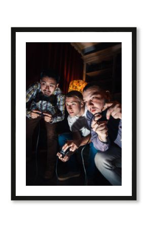Excited and tensioned video game tourney, company of three high concentrated young guys play with joysticks at home. Competition, fun entertainment concept