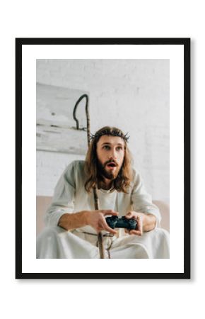 emotional Jesus with wooden staff playing video game by joystick on sofa at home