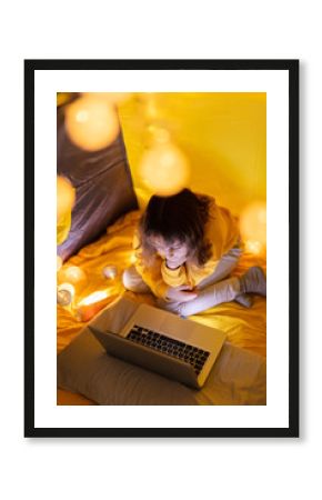 Young girl student using laptop for homeschooling in a home made tent with glowing light balls.