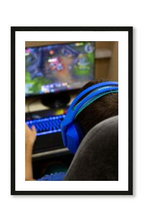 Back view of male gamer wearing blue headphones playing computer game at home. Setup includes gaming monitor, mechanical keyboard with colorful lighting, comfortable chair