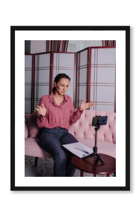 a woman psychologist in a pink shirt is sitting on the couch and conducting an online consultation using her phone