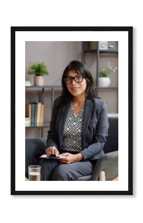 Vertical medium shot of young adult woman working as psychologist sitting on chair in her office holding clipboard with notebook looking at camera