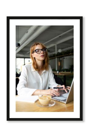 Young woman with with red hair in eyeglasses and white shirt sitting at the table with a laptop