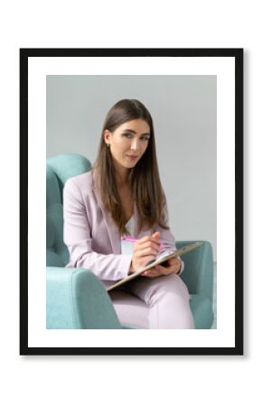 Middle-aged psychologist woman in a beautiful modern suit. Sitting in a comfortable chair for a consultation. Makes notes on clipboard.