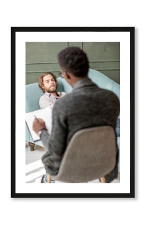 Young caucasian man visiting an african psychologist lying on the comfortable couch during psychological session in the green office