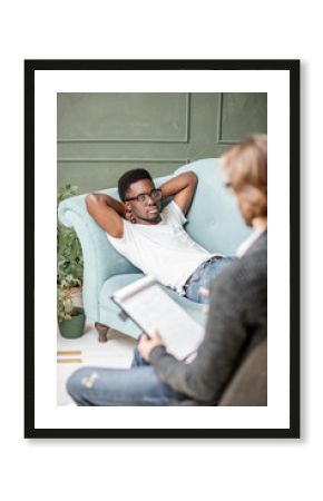 Young afro ethnicity man having a serious conversation with psychologist lying on the comfortable couch during psychological session in the office