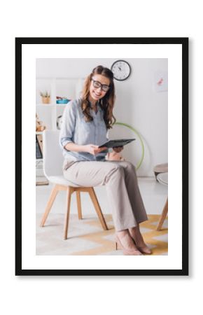 smiling child psychologist with clipboard sitting on chair in office and looking at camera