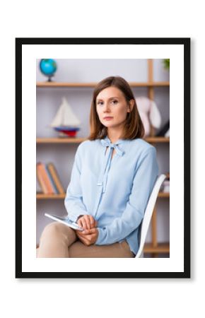 Female psychologist with digital tablet looking at camera while sitting on chair on blurred background