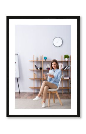 Full length of smiling psychologist with digital tablet looking at camera while sitting on chair in office