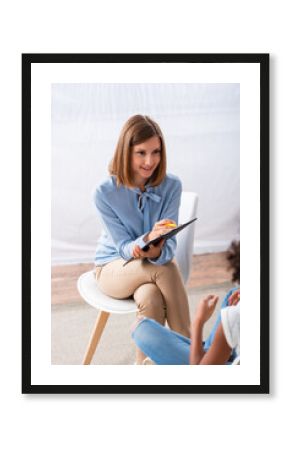Smiling psychologist with clipboard looking at african american girl during consultation on blurred foreground