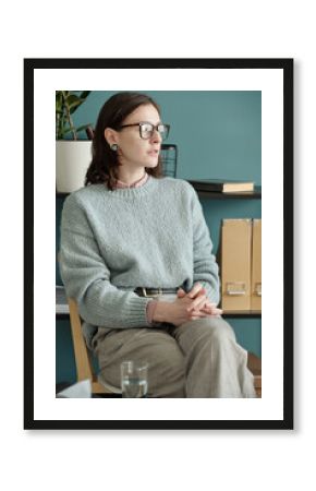 Portrait of woman sitting with hands clasped during therapy session, wearing glasses and looking thoughtfully to side in professional office setting
