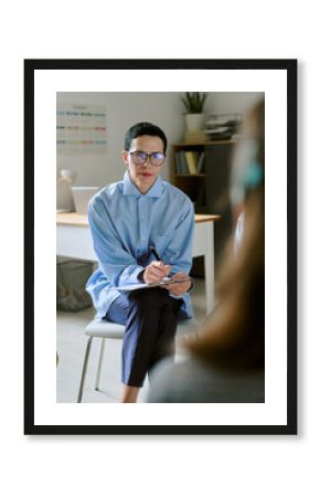 Middle aged Caucasian woman wearing glasses sitting on chair holding clipboard and pen listening attentively to unseen in office setting during consultation session