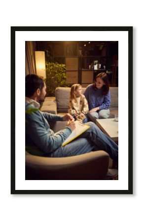 Happy mother and her little daughter sitting on sofa during their therapy session with psychologist taking notes. Maintaining healthy space for kids growth and supportive communication. Psychology