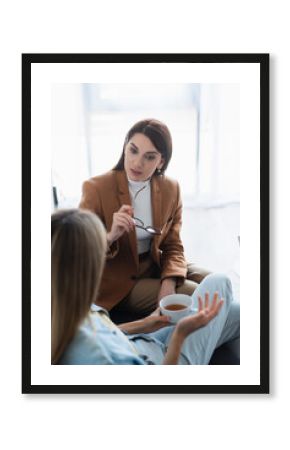 psychologist holding eyeglasses while talking to blurred woman with cup of tea