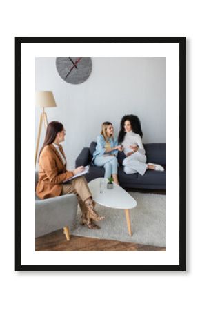 psychologist with clipboard listening to smiling interracial lesbian couple sitting on couch in consulting room