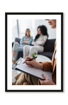 psychologist writing on clipboard near blurred multiethnic lesbians sitting on couch