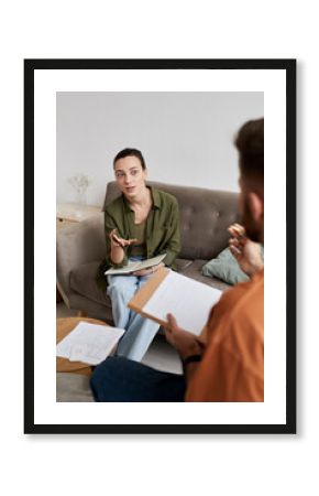 Confident tutor with copybook discussing new subject points with student while sitting on couch in front of him at home lesson