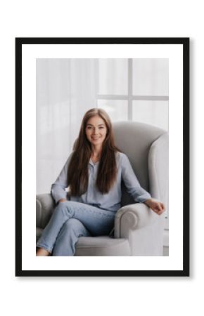 Satisfied young woman in blue blouse and jeans sitting in chair broad smiles. Blonde confident female psychologist waiting for client. Mental health. Successful caucasian girl relaxing at home. Model