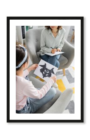 High angle view of blurred preteen child holding Rorschach test near psychologist with clipboard in consulting room.