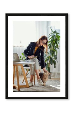 Young woman using laptop and mobile phone stroking a pet cat at workplace at home, remote work