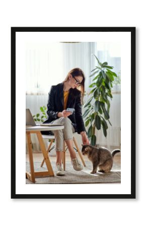 Young woman using laptop and mobile phone stroking a pet cat at workplace at home, remote work
